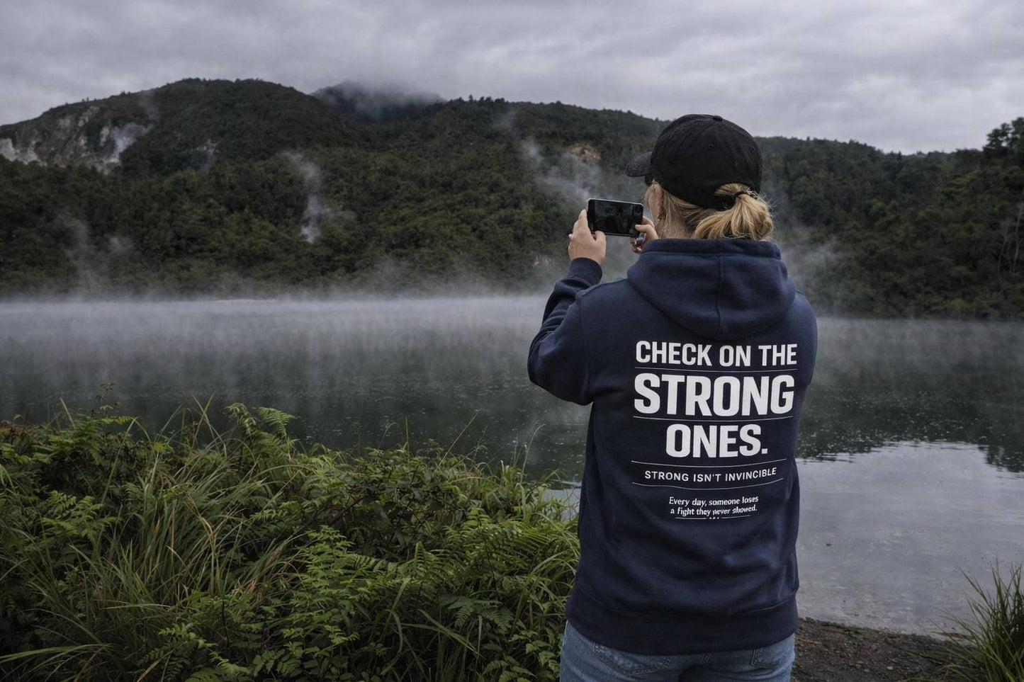 Person taking a photo of a lake with mountains in the background, wearing a hoodie with text.