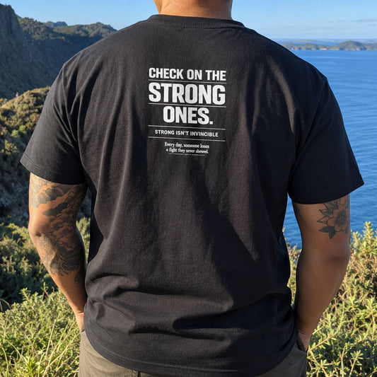 A man wearing a black t-shirt with a white printed quote on the back, standing outdoors on a cliffside overlooking the ocean.