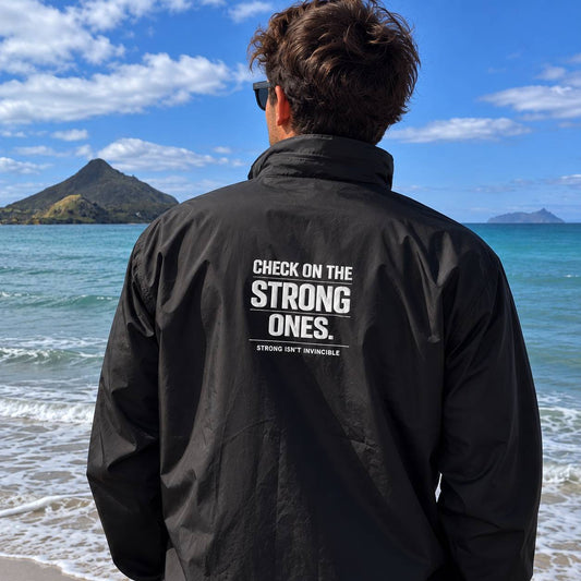 A man viewed from behind wearing a black windbreaker jacket with 'CHECK ON THE STRONG ONES.' printed in white on the back, standing on a beach with the ocean and mountains in the background.