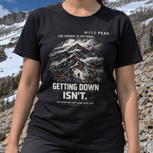 A person outdoors in a rocky mountain setting wearing a black graphic T-shirt. The shirt features a snow and rock mountain illustration with orange highlights and white text reading 'WYLD PEAK' and 'GETTING DOWN ISN’T.