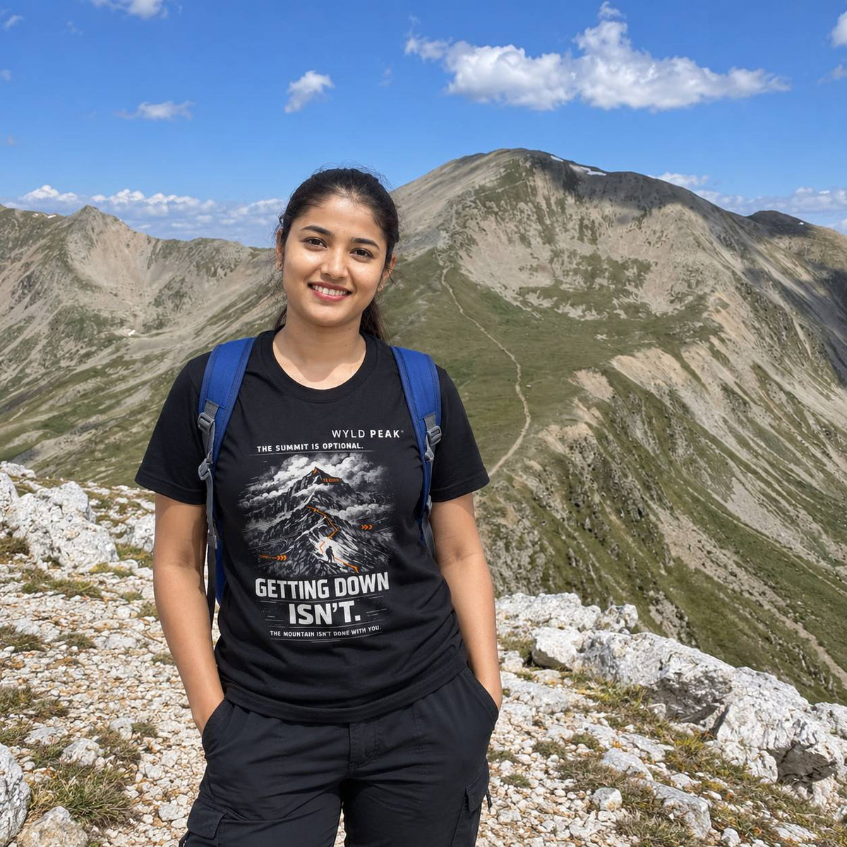 A smiling woman stands on a mountain peak wearing a black graphic t-shirt with a blue backpack, set against a backdrop of rugged mountains and a blue sky.