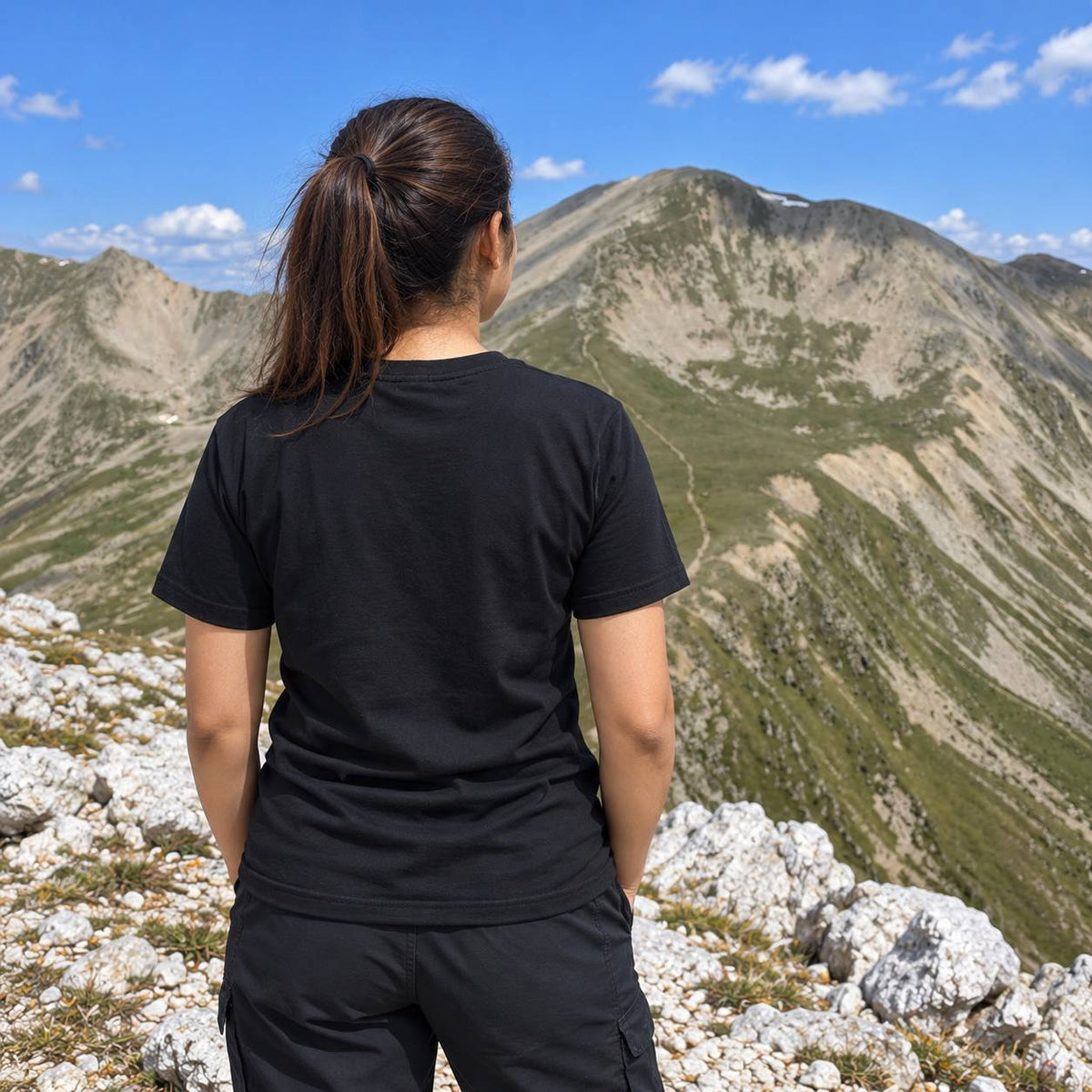 A woman seen from behind wearing a black short-sleeve t-shirt and black cargo pants, standing on a rocky mountain peak with a vast green and gray mountain range and blue sky in the background.