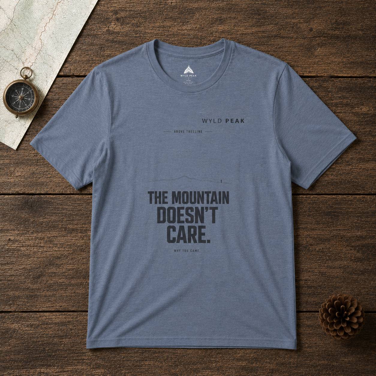 Blue t-shirt with 'The Mountain Doesn't Care' text on a wooden surface with a compass and pine cone.