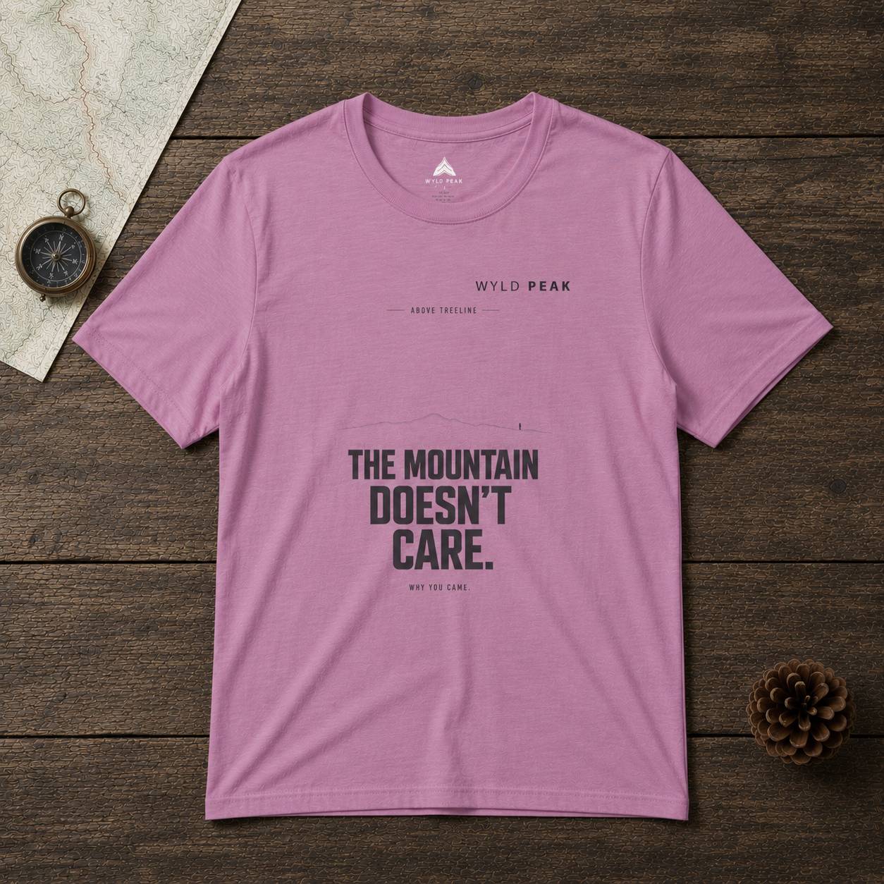 Pink t-shirt with 'The Mountain Doesn't Care' text on a wooden surface with a compass and pine cone.