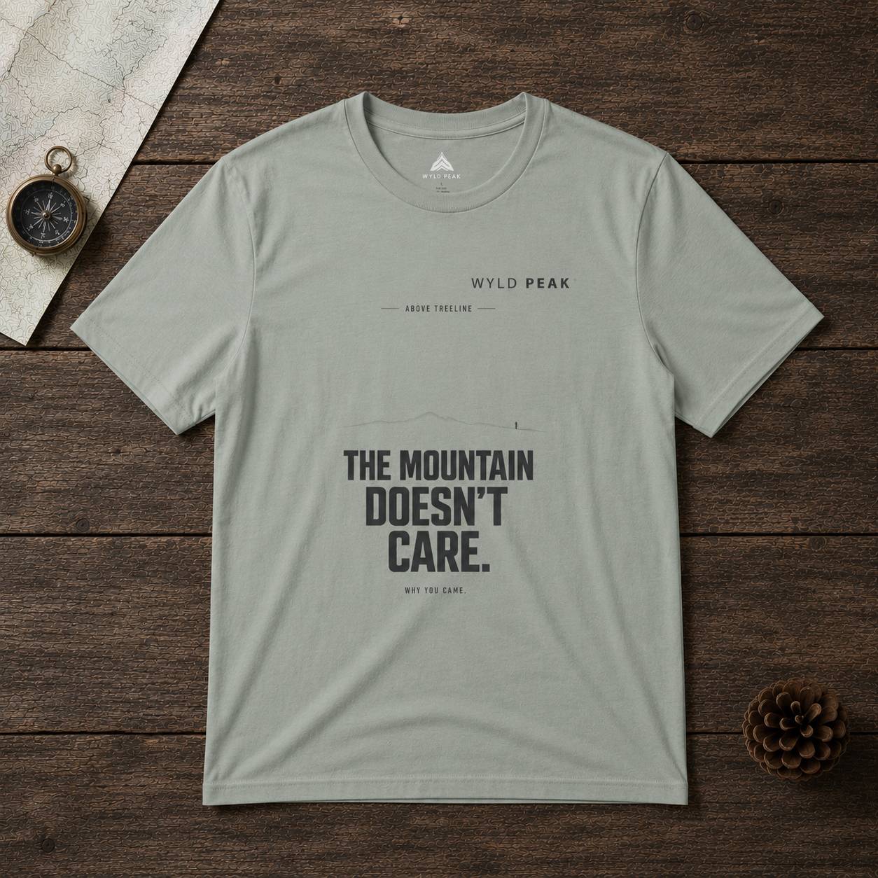 Gray t-shirt with 'The Mountain Doesn't Care' text on a wooden surface with compass and map.
