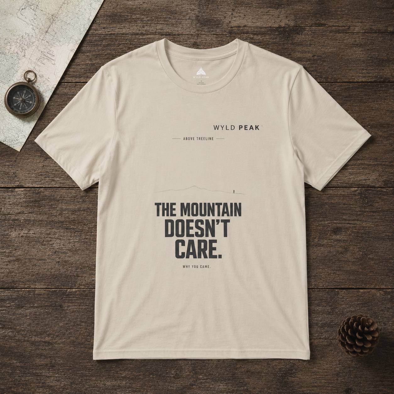 Beige t-shirt with text on a wooden surface with a compass and pine cone.