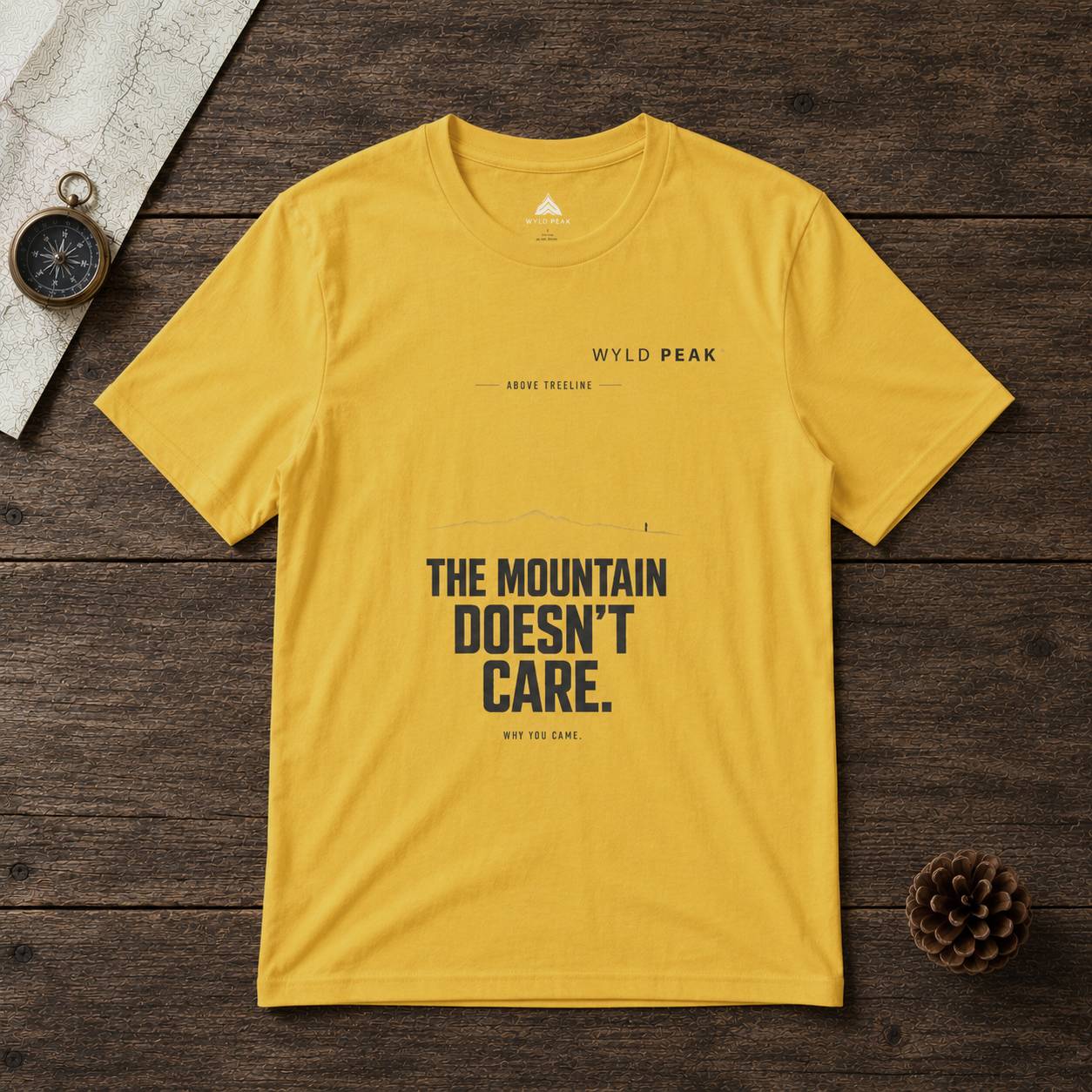 Yellow t-shirt with 'The Mountain Doesn't Care' text on a wooden surface with a compass and pine cone.