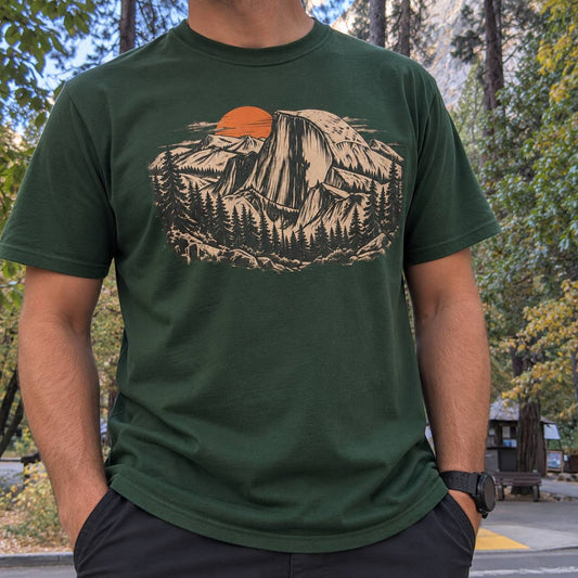 A man wearing a dark green t-shirt with a graphic print of a mountain sunset in beige and orange, photographed outdoors in a park setting.
