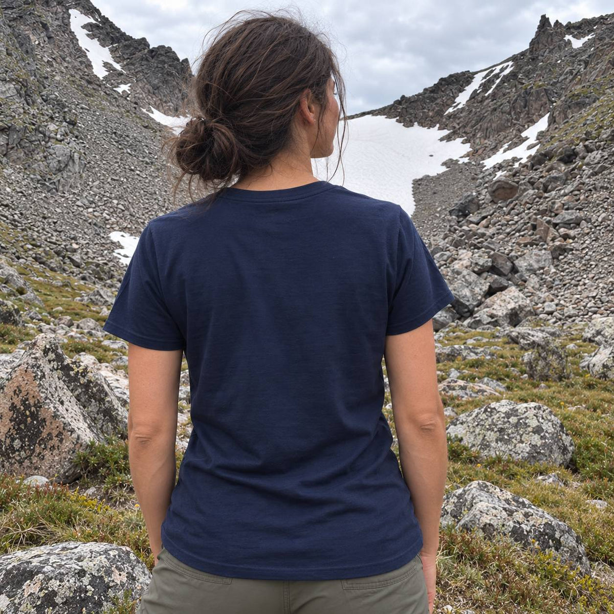 A woman viewed from behind wearing a navy blue short-sleeve crewneck t-shirt, standing in a rocky mountain landscape with patches of snow under an overcast sky.