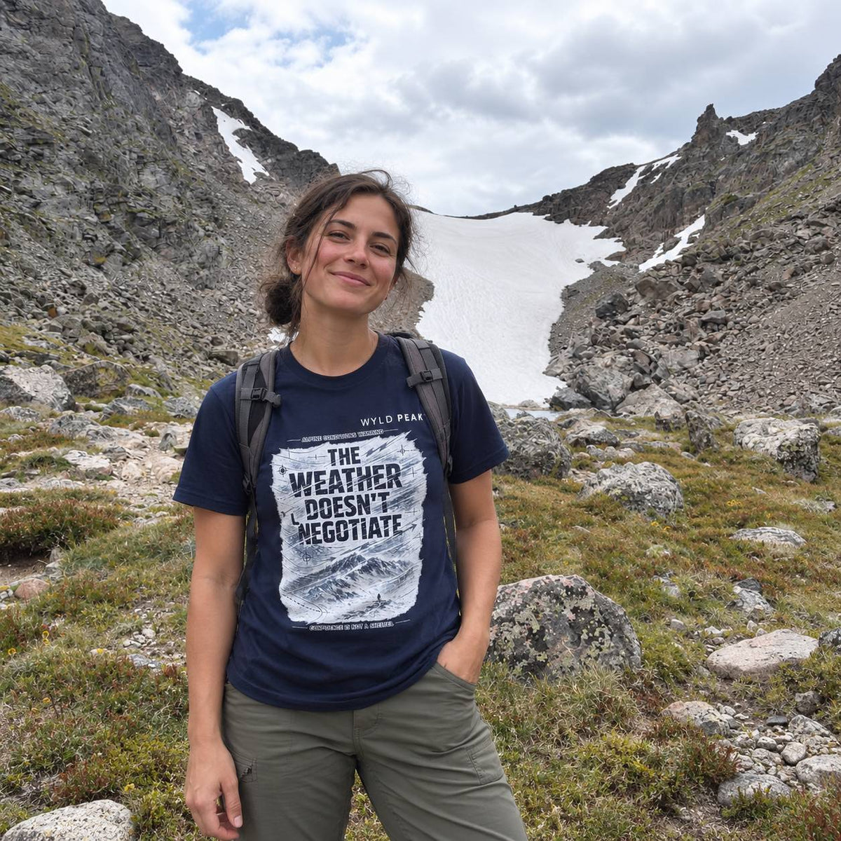 A woman hiking in a rocky mountain pass with snow patches in the background, wearing a navy blue graphic t-shirt that reads 'THE WEATHER DOESN'T NEGOTIATE' and olive green pants.