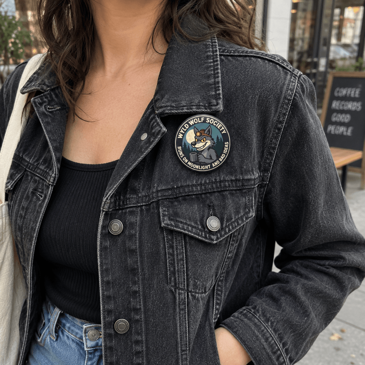A person wearing a dark washed denim jacket with a circular 'Wild Wolf Society' pin on the chest, paired with a black ribbed top and blue jeans in an outdoor urban setting.