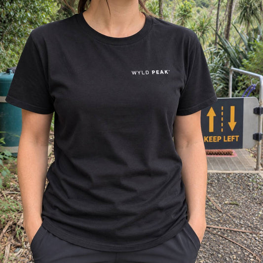 A woman wearing a black crewneck t-shirt with 'WYLD PEAK' printed in white on the chest, standing outdoors in a forest setting.