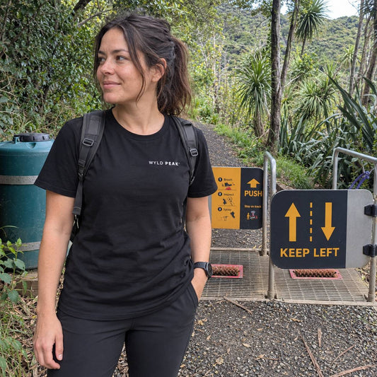 A woman wearing a black t-shirt with 'WYLD PEAK' printed on the chest stands at a trail entrance with lush green forest in the background.