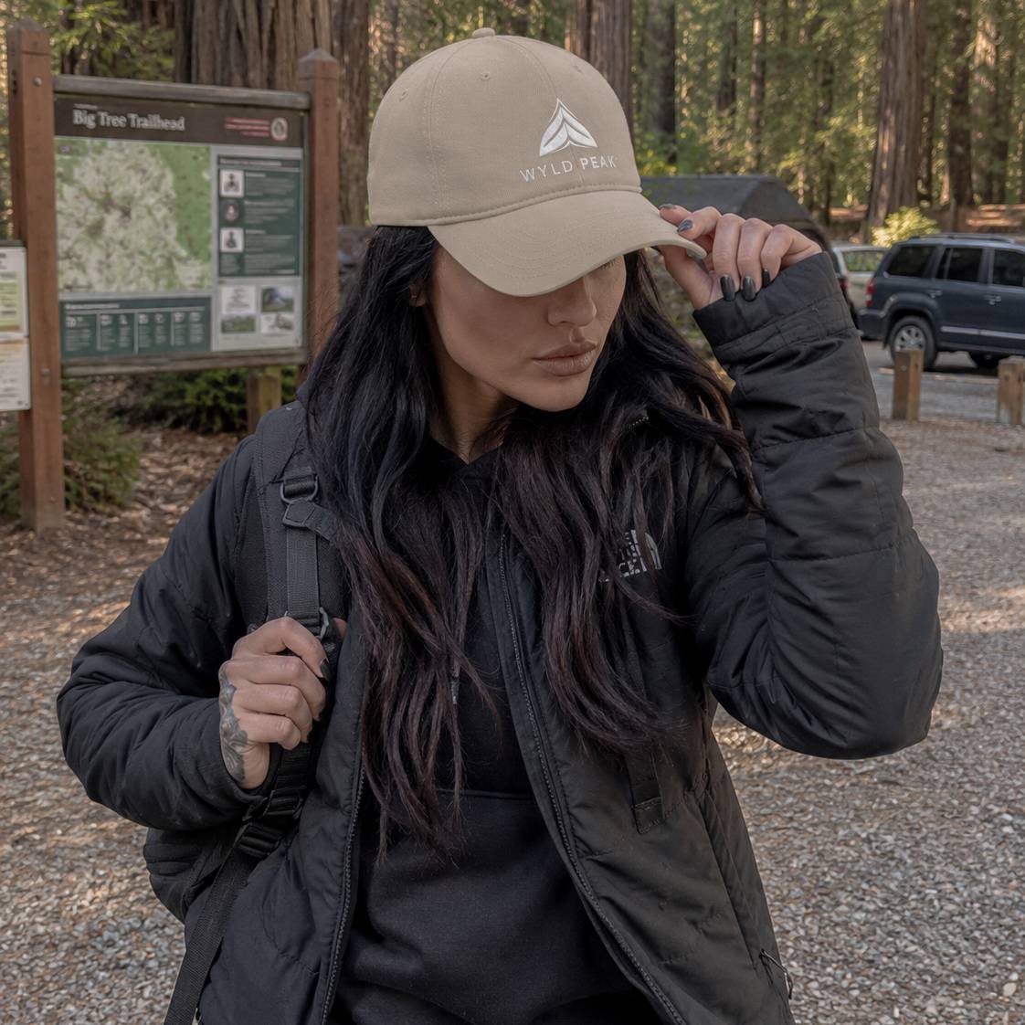 Woman wearing a beige cap with a logo, standing in a forested area with a trailhead sign in the background.