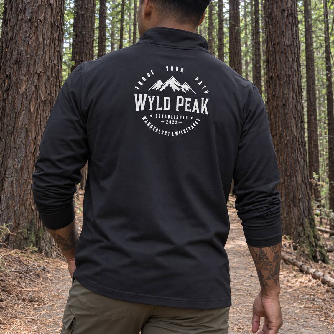 A man viewed from behind wearing a black long-sleeve quarter-zip pullover with a white 'Wyld Peak' mountain graphic on the back, standing on a forest path.