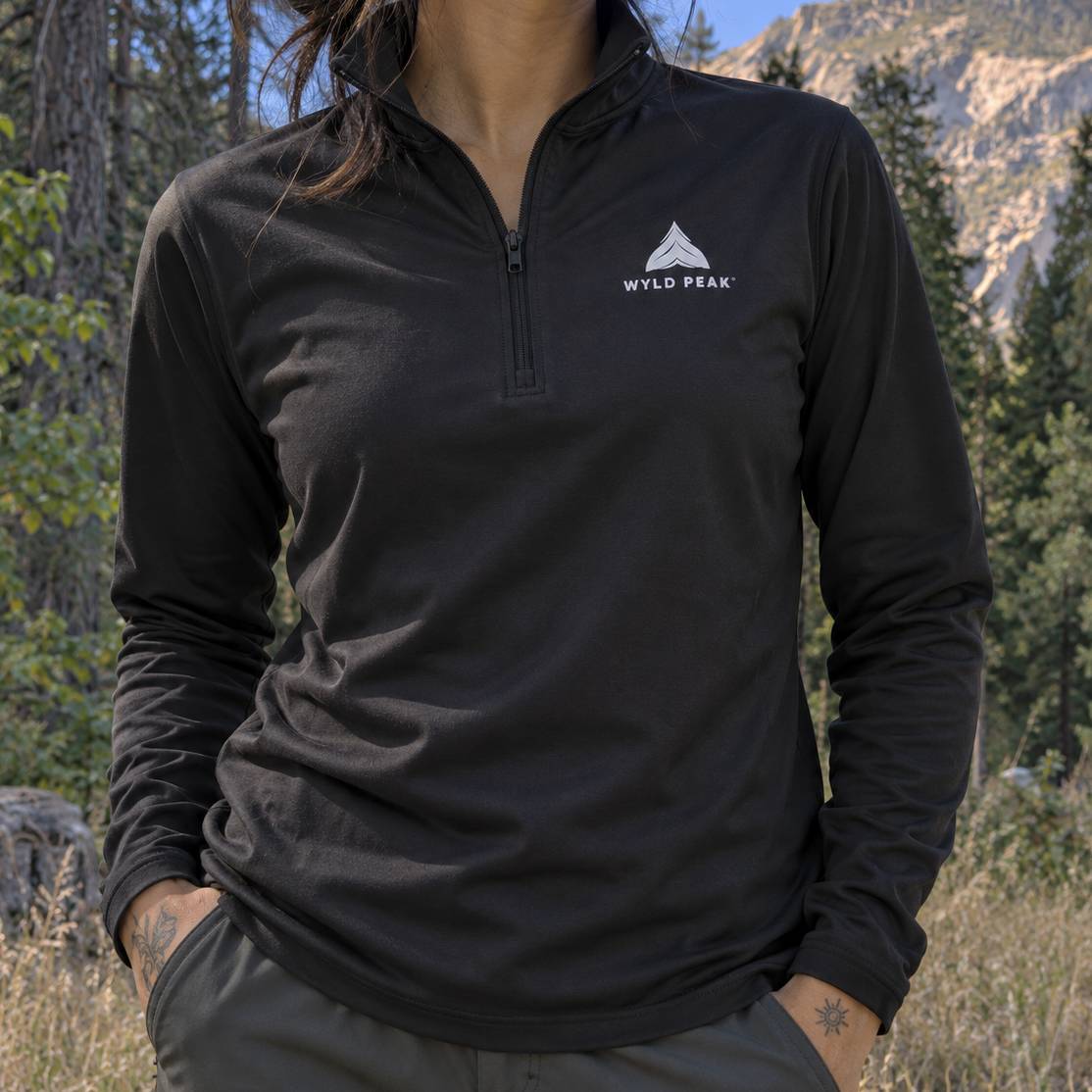 A woman wearing a black long-sleeve quarter-zip pullover with a white Wild Peak logo on the left chest, photographed outdoors in a mountain and forest setting.