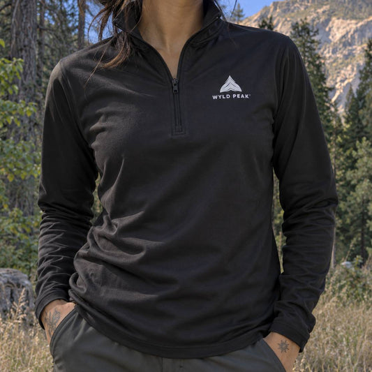 A woman wearing a black long-sleeve quarter-zip pullover with a white Wild Peak logo on the left chest, photographed outdoors in a mountain and forest setting.