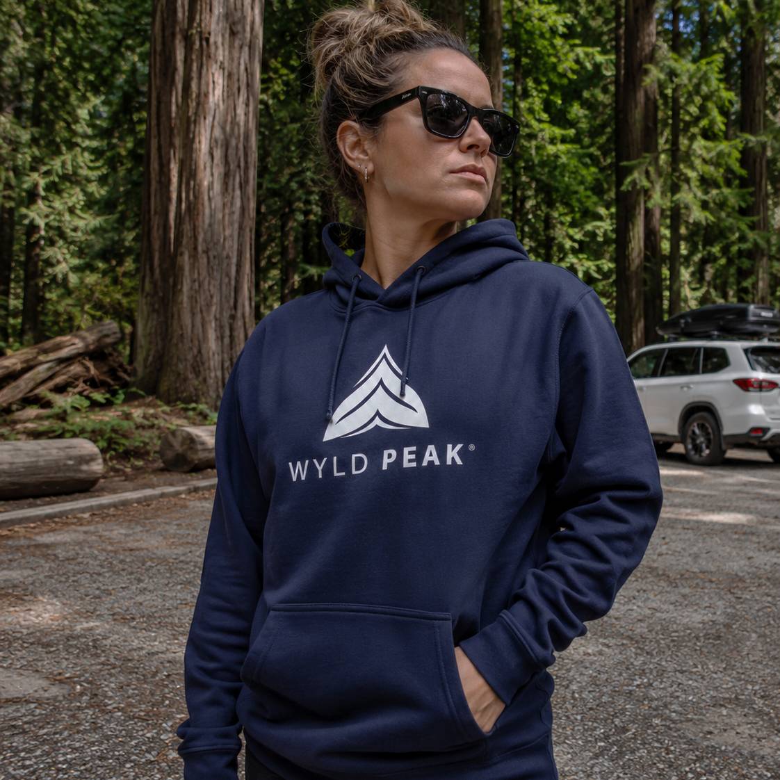 Woman wearing a navy blue hoodie with 'Wyld Peak' logo in a forest setting