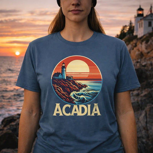 Person wearing a blue t-shirt with a lighthouse graphic and 'Acadia' text, standing in front of a sunset over water.