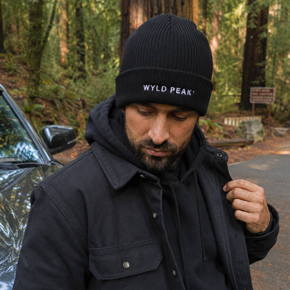 Man wearing a black beanie with 'WYLD PEAK' logo in a forest setting