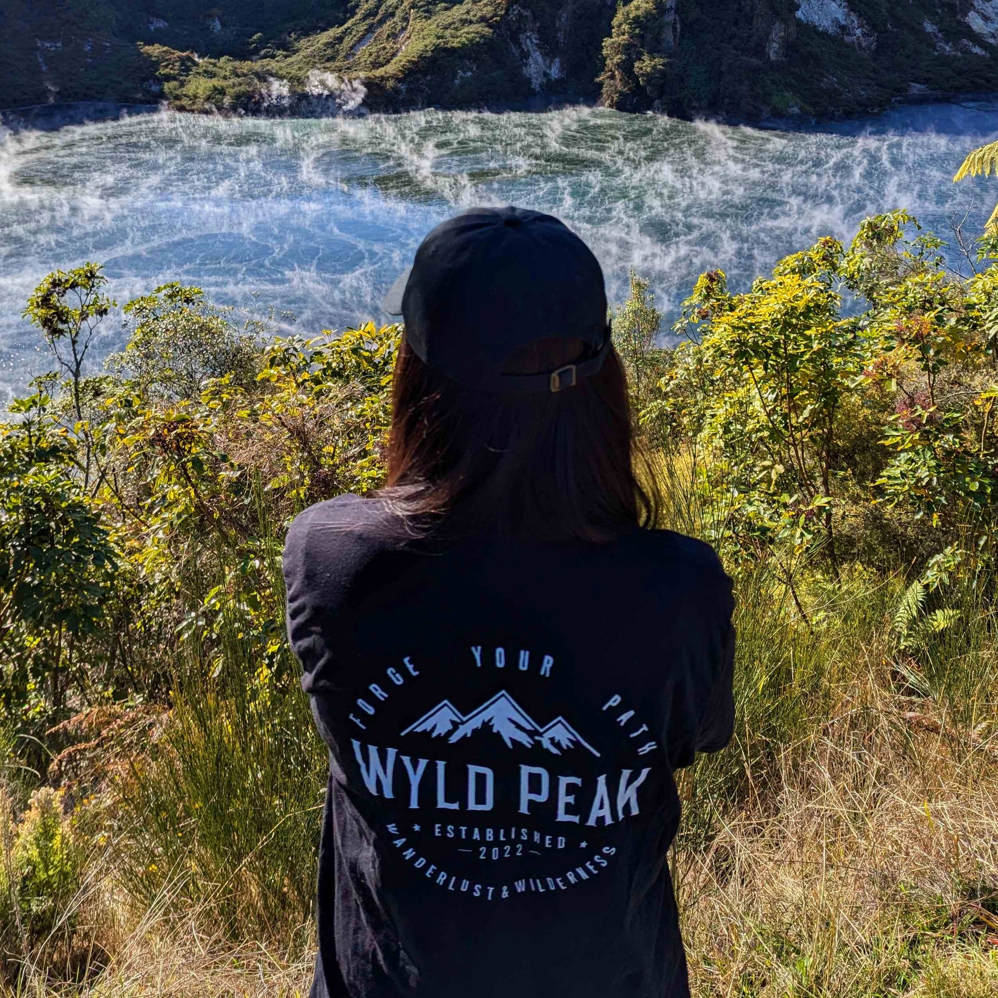 Person wearing a black 'Wyld Peak' shirt overlooking a scenic landscape with water and mountains.