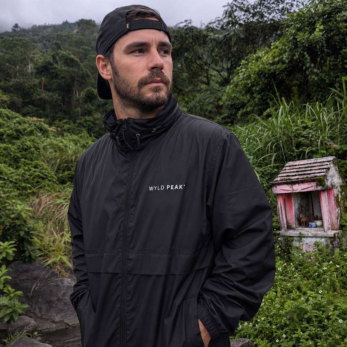 A man wearing a black waterproof jacket with 'WILD PEAK' printed in white on the chest, standing outdoors in a lush green mountain environment.