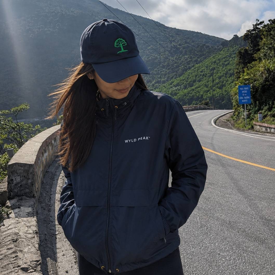 A woman wearing a navy blue zip-up jacket and matching baseball cap stands on a mountain road. The jacket has a small white 'WYLD PEAK.