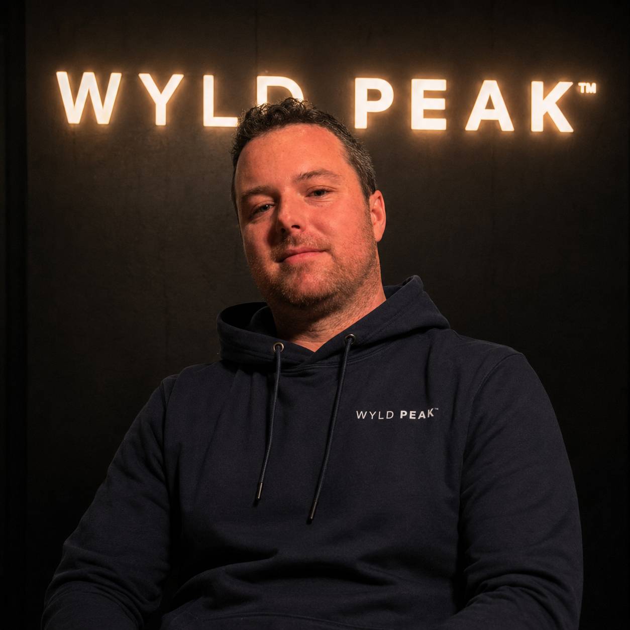 Man wearing a dark hoodie with 'WYLD PEAK' logo in front of a dark background with 'WYLD PEAK' branding.