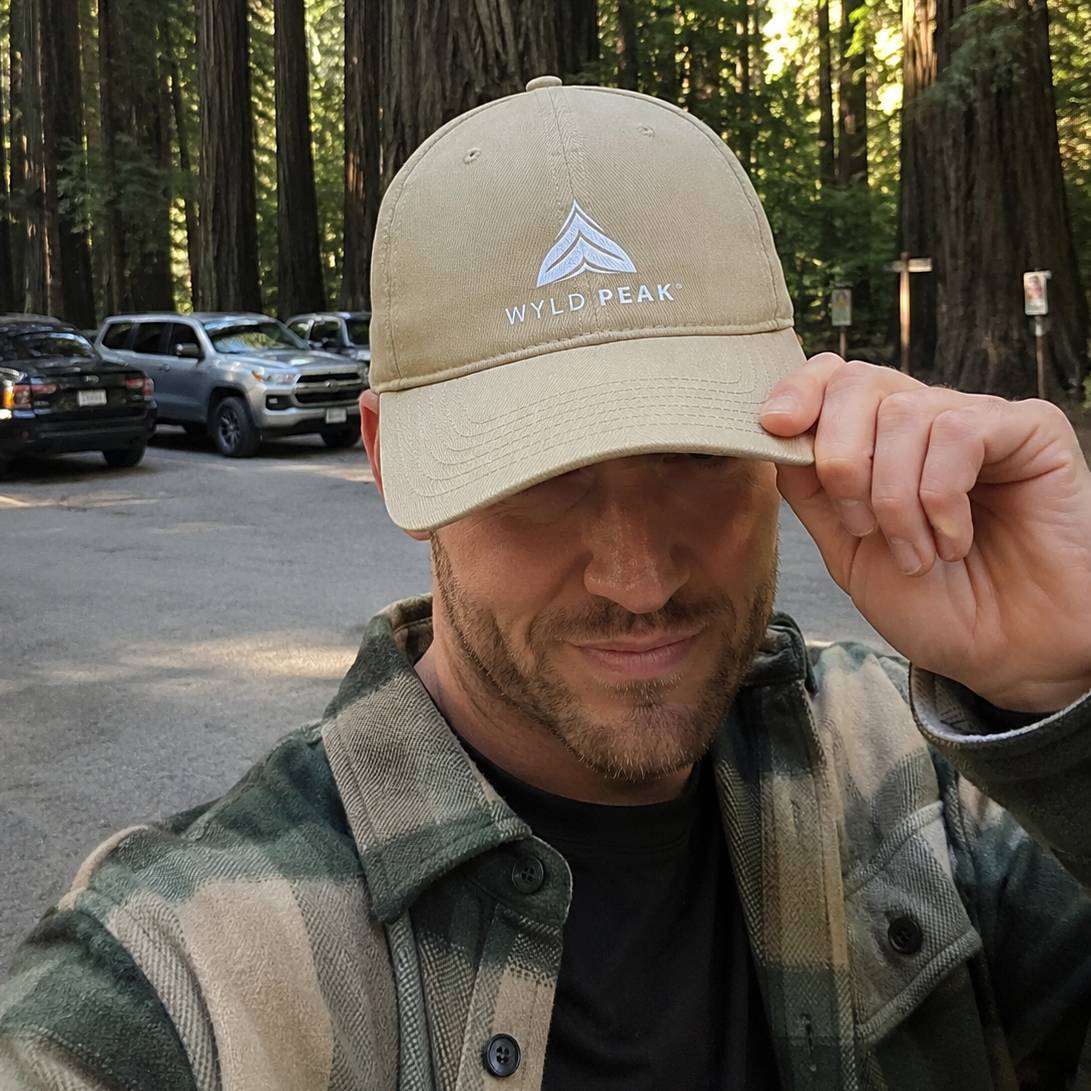 Man holding a beige cap with 'Wyld Peak' logo in a forest setting