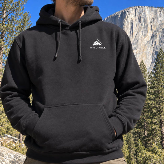 A man wearing a black pullover hoodie with a white 'WYLD PEAK' logo on the left chest, photographed outdoors against a mountain and pine tree background.