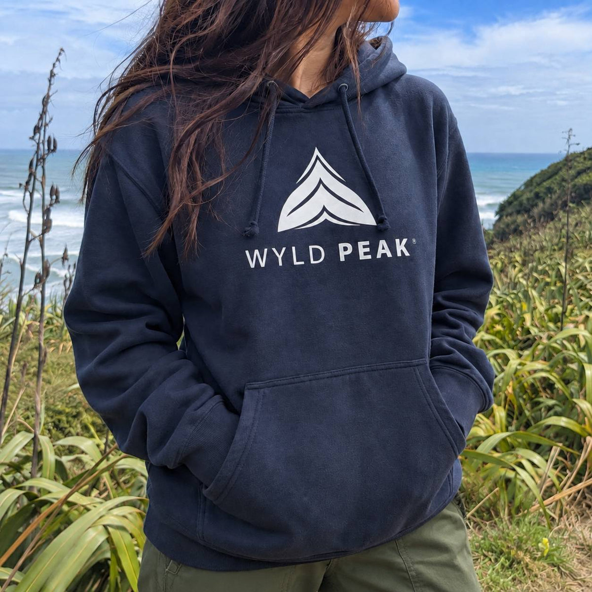 A person wearing a navy pullover hoodie with a white 'WYLD PEAK' logo and mountain graphic on the chest, standing outdoors on a grassy cliffside with the ocean and sky in the background.