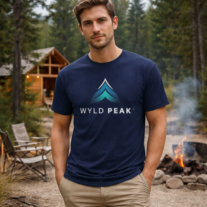 Man wearing a navy blue 'Wyld Peak' t-shirt in a forest setting with a cabin and campfire.