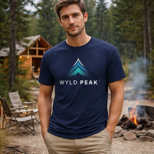 Man wearing a navy blue 'Wyld Peak' t-shirt in a forest setting with a cabin and campfire.
