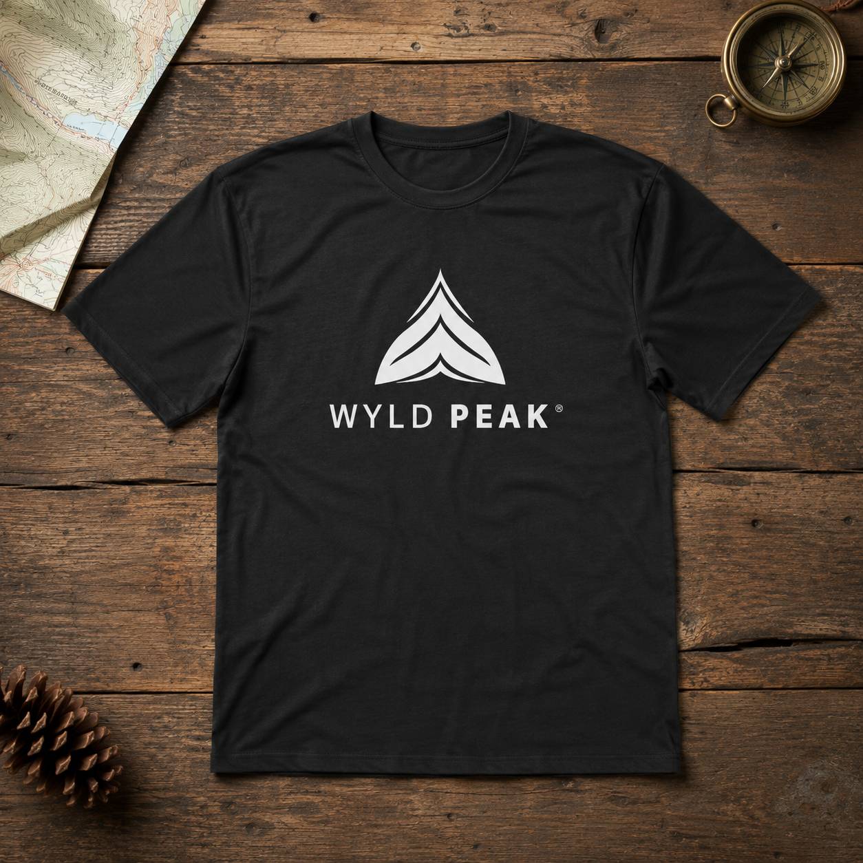 A black short-sleeve t-shirt with a white graphic logo reading 'WYLD PEAK' and a mountain-style icon, laid flat on a rustic wooden surface with a map, pinecone, and compass as props.