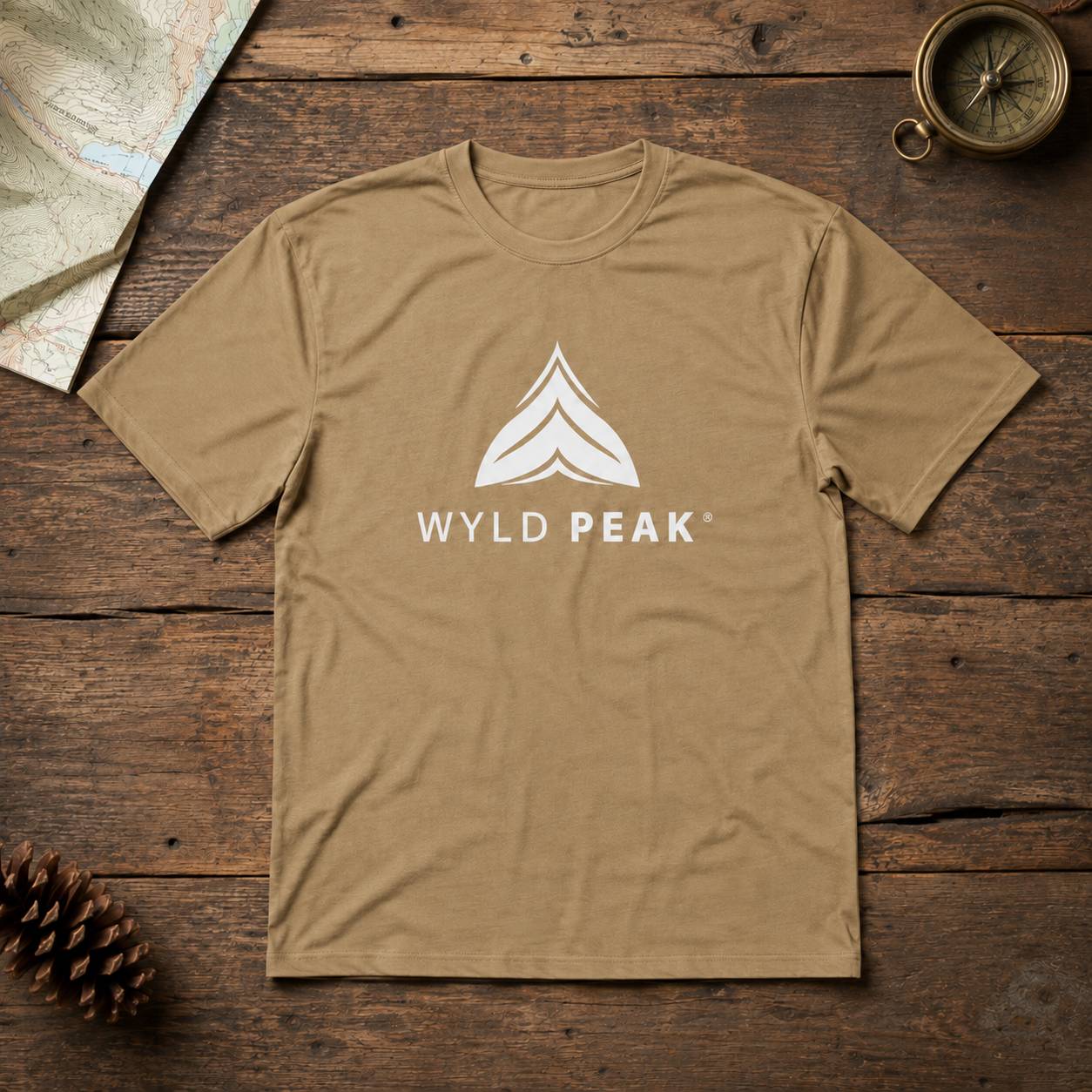 A tan crewneck t-shirt with a white 'WYLD PEAK' logo and mountain graphic on the chest, laid flat on a rustic dark wood background with a map, pinecone, and compass as props.