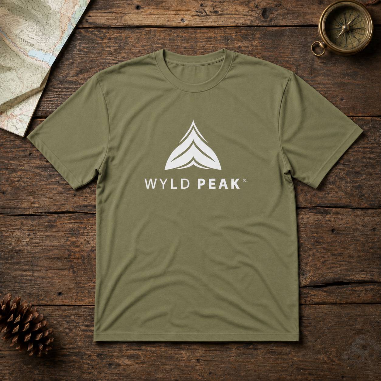 An olive green crewneck t-shirt with a white 'WYLD PEAK' logo and mountain graphic on the chest, laid flat on a rustic dark wood background with a map, pinecone, and compass as props.
