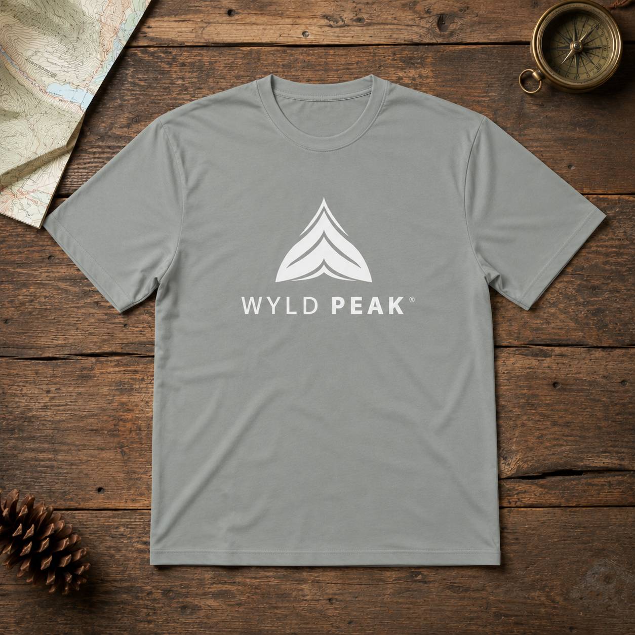 A light gray crewneck t-shirt with a white 'WYLD PEAK' logo and mountain graphic on the chest, laid flat on a rustic dark wood background with a map, pinecone, and compass as props.