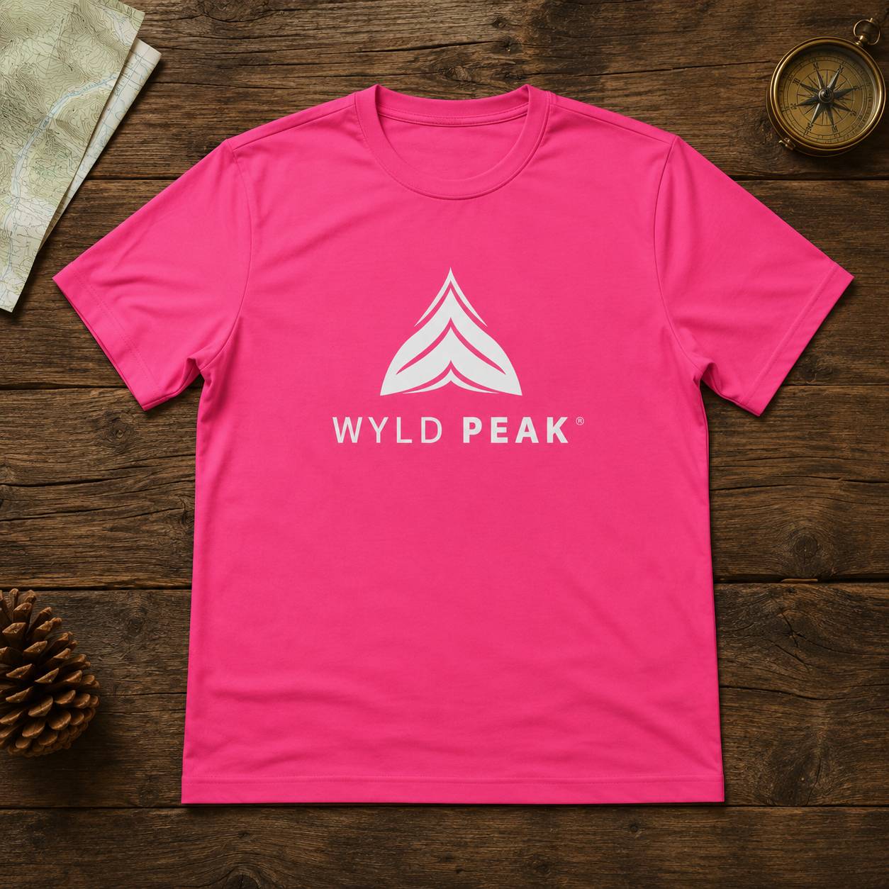 A bright pink crewneck t-shirt with a white 'WYLD PEAK' logo and mountain graphic on the chest, laid flat on a dark wood background with outdoor-themed props.