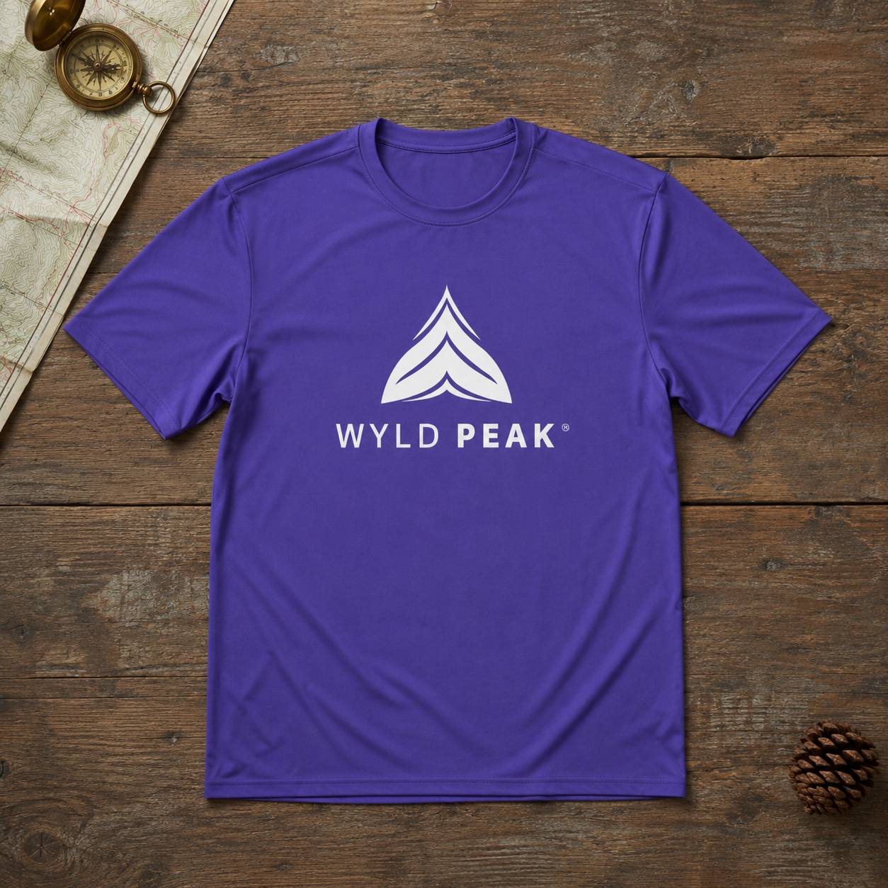 A purple short-sleeve crewneck t-shirt with a white 'WYLD PEAK' logo printed on the chest, laid flat on a rustic wooden surface with a compass and pinecone props.