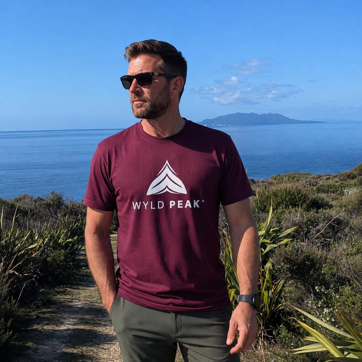 A man wearing a burgundy t-shirt with a white 'WYLD PEAK' logo stands outdoors on a path with a scenic ocean and mountain background.