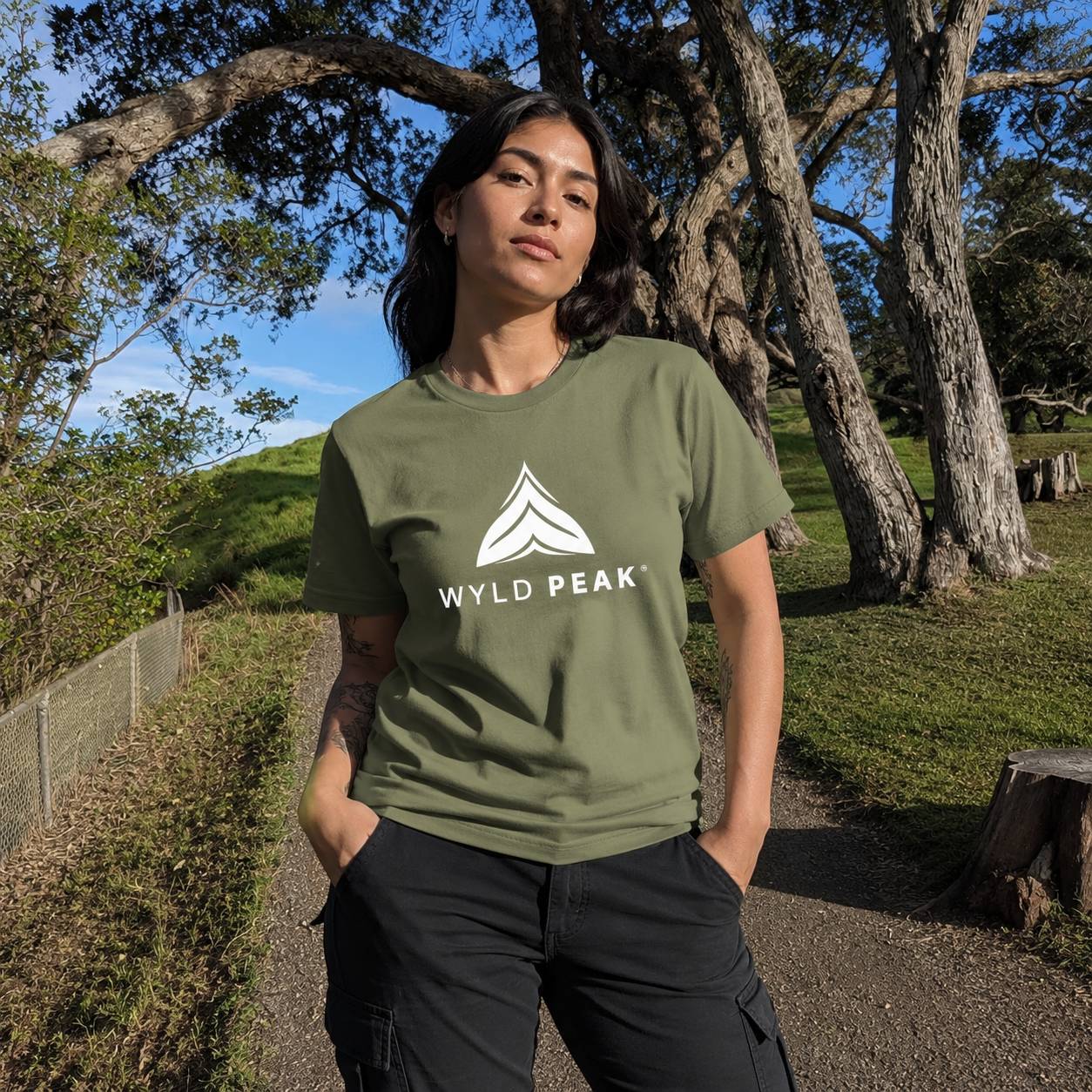 A woman wearing an olive green t-shirt with a white geometric logo and the text 'WYLD PEAK' printed on the chest, standing outdoors in a park with trees and grass.