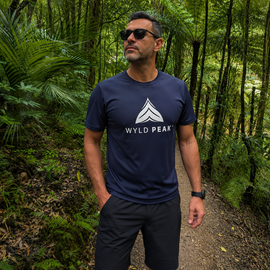 A man wearing a navy blue t-shirt with a white geometric logo and the text 'WYLD PEAK' stands on a forest path wearing dark shorts and sunglasses.