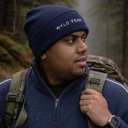 Man wearing a blue 'Wyld Peak' beanie and carrying a backpack in a forest setting