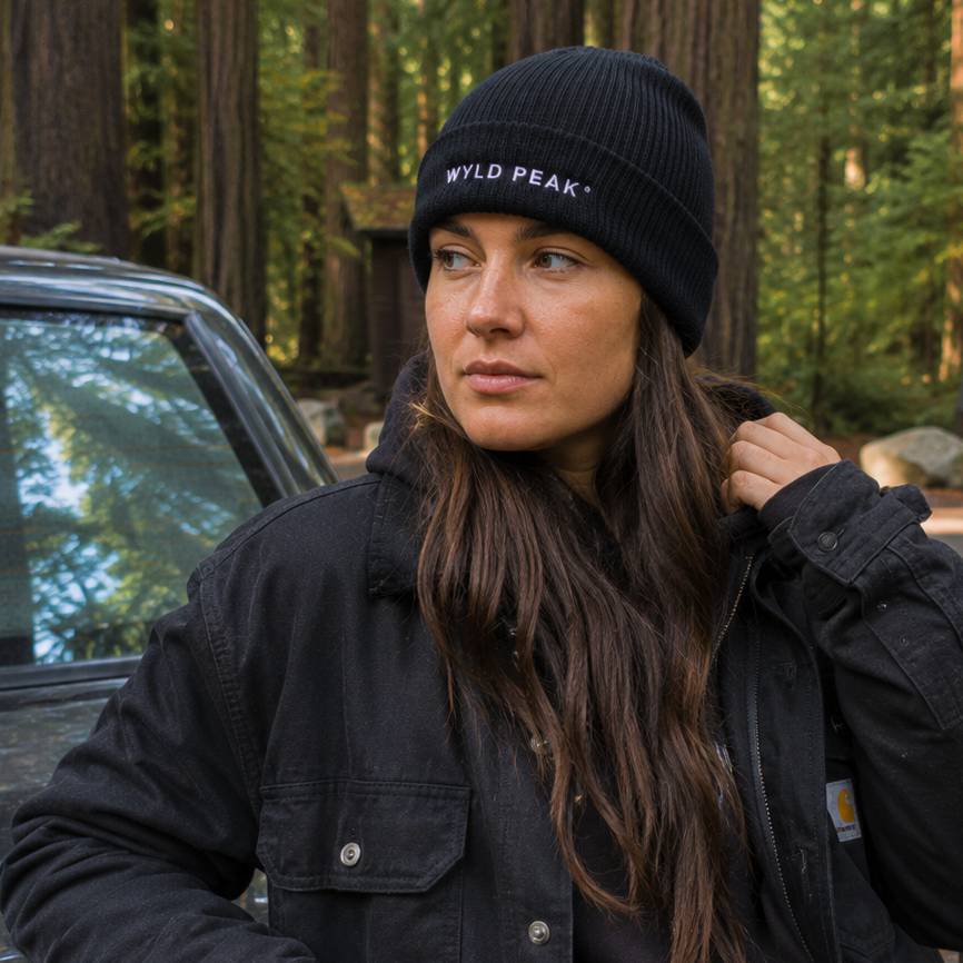 Woman wearing a black jacket and 'WYLD PEAK' beanie in a forest setting
