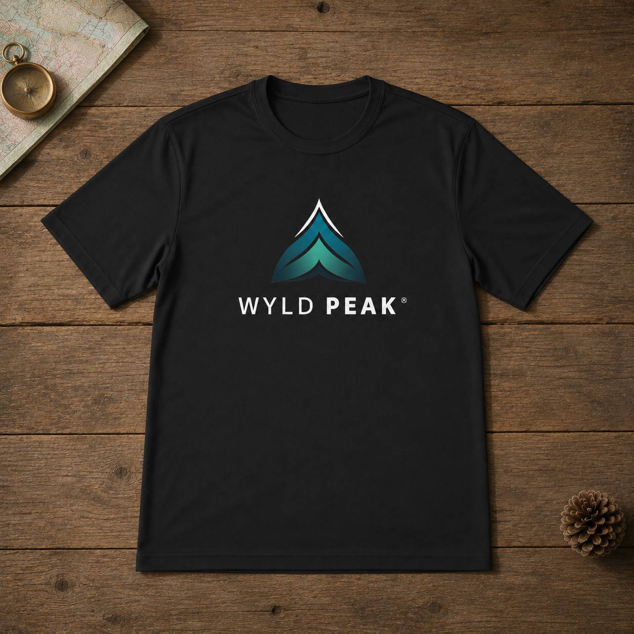 A black short-sleeve crewneck t-shirt featuring a stylized mountain logo in teal and white with the text 'WYLD PEAK' underneath, laid flat on a rustic wooden background with a map, brass compass, and pinecone as props.