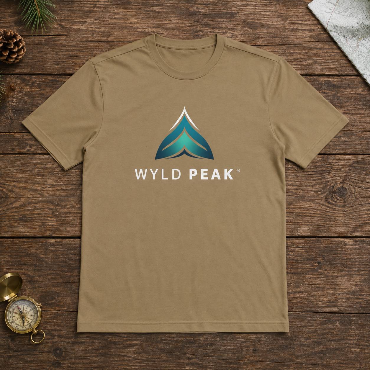 A tan crewneck t-shirt with a blue and white 'WYLD PEAK' logo on the chest, laid flat on a dark wood background with decorative props.