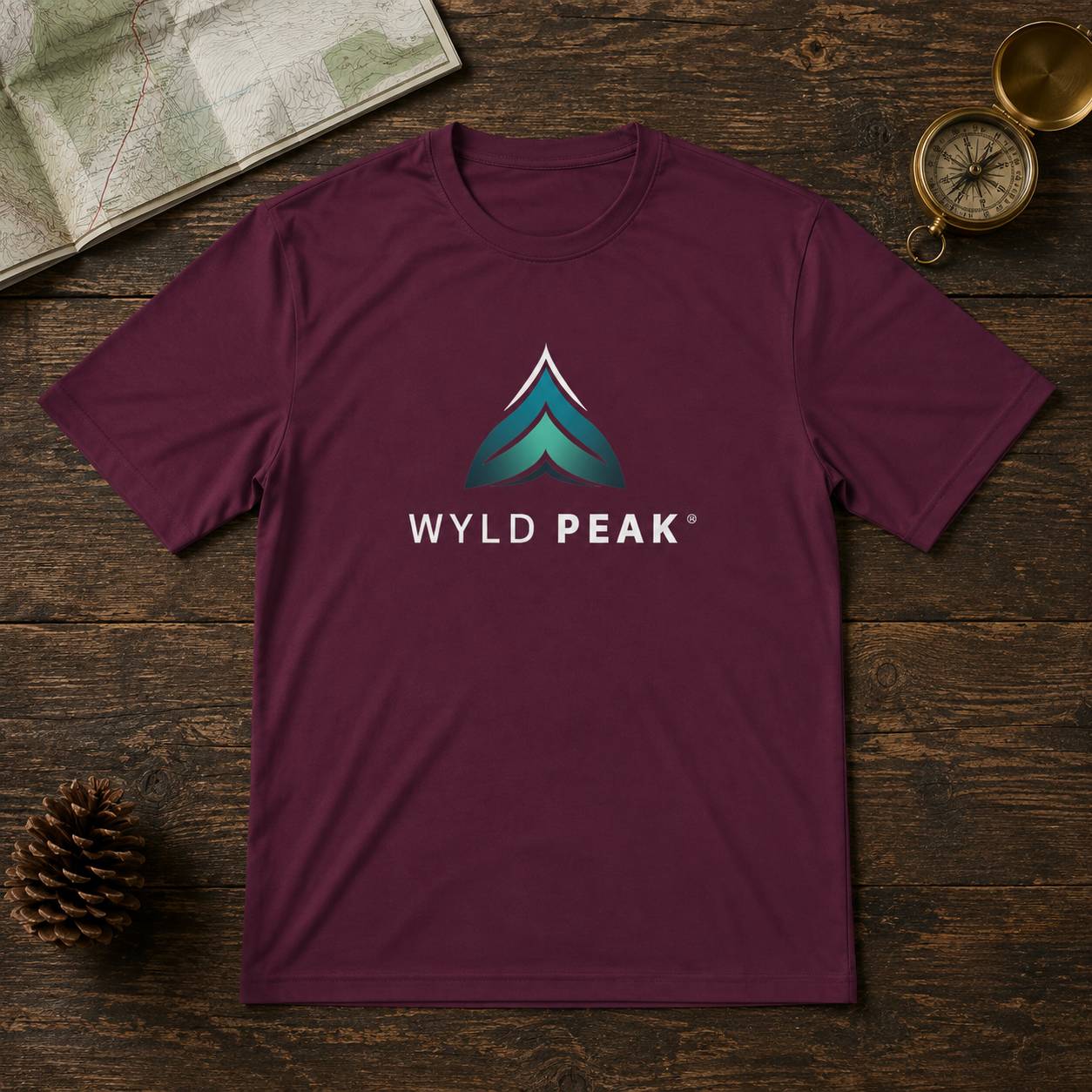 A maroon short-sleeve crewneck t-shirt featuring a teal and white 'WYLD PEAK' mountain logo on the chest, laid flat on a dark wood background with a map, pinecone, and compass as props.