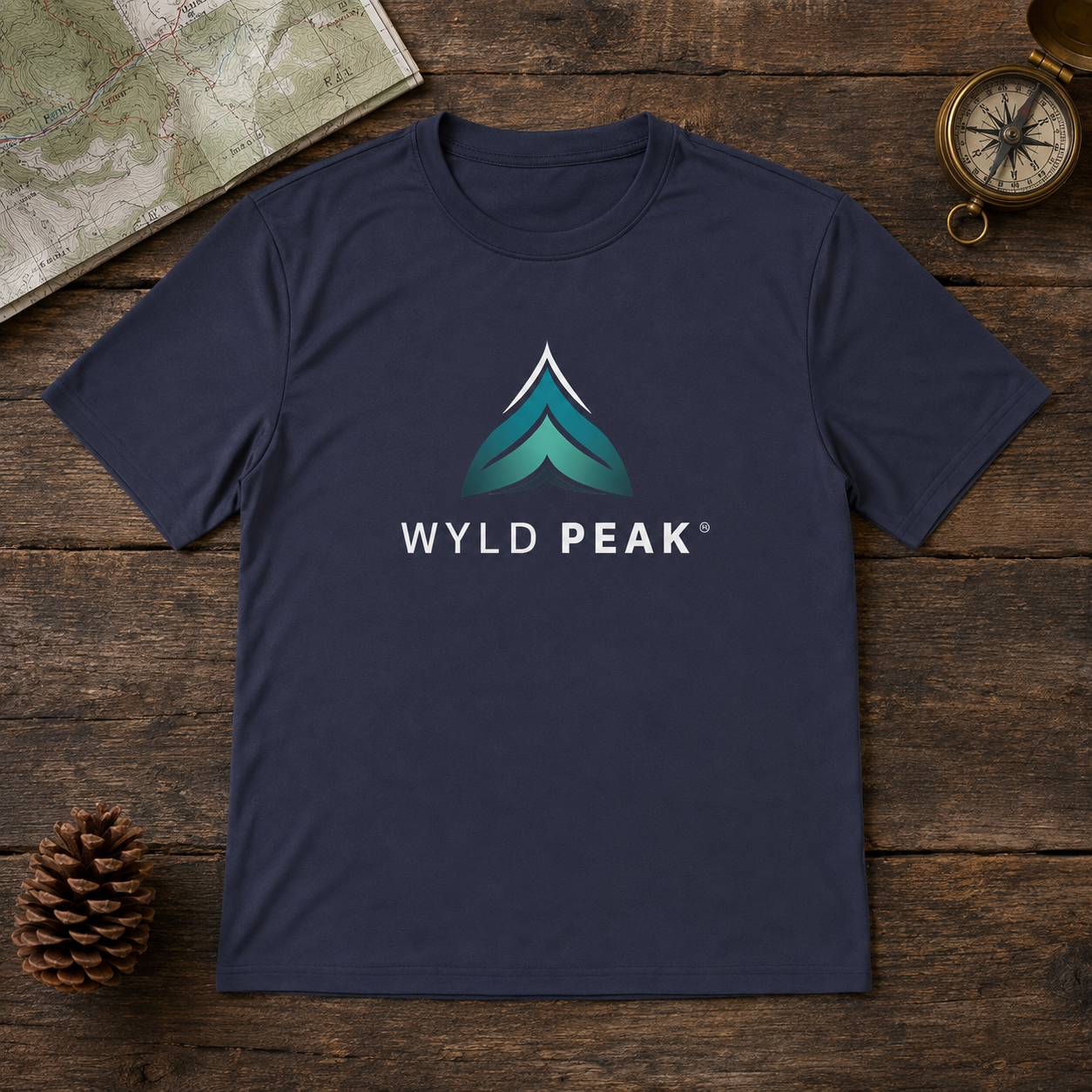 A navy blue crewneck t-shirt featuring a stylized mountain logo in teal and white with the text 'WYLD PEAK' printed on the chest. The t-shirt is laid flat on a rustic dark wood background, surrounded by decorative props including a vintage map, a pinecone, and a compass.