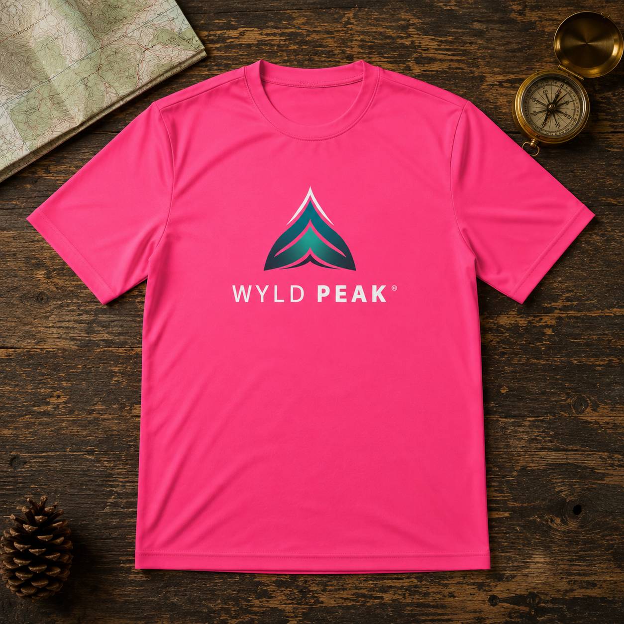 A bright pink crewneck t-shirt featuring a stylized mountain logo and the text 'WYLD PEAK' on the chest, laid flat on a dark rustic wooden background with a map, pinecone, and compass as props.