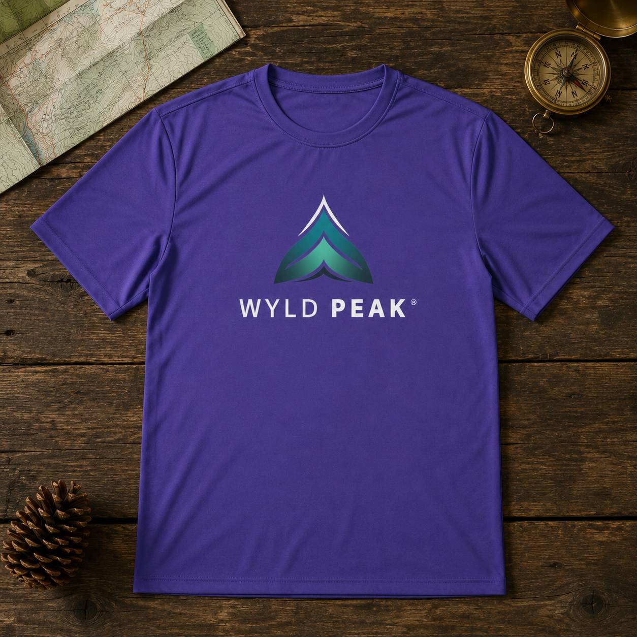 A purple crewneck t-shirt featuring a green and blue mountain graphic with the text 'WYLD PEAK' printed on the chest, laid flat on a rustic dark wood background with a map, pinecone, and compass as props.